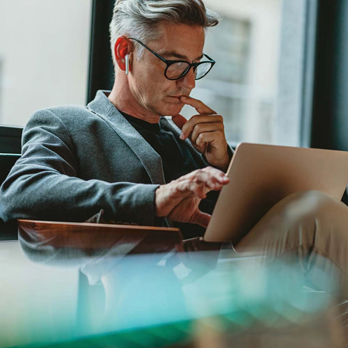 gray-haired man works on laptop in front of large window